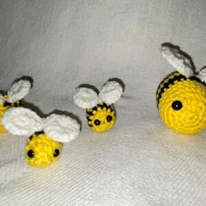 May include: Four handmade crochet bees in various sizes. The bees are yellow with black stripes and white wings. They have black eyes and are arranged on a white textured surface.