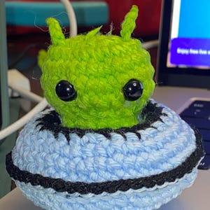 May include: A handmade crochet alien figure with a green head and large black eyes. The alien sits atop a light blue and black crocheted UFO. The craft has a black trim and a white accent. The alien has two small antennae.