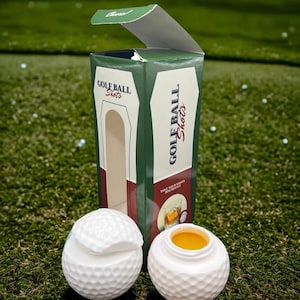 May include: Two white golf ball shot glasses with a green and brown box that says "Golf Ball Shots" on the side. The box is open and the shot glasses are on a green grassy surface.