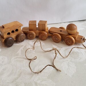 May include: A wooden toy train set with three connected cars. The engine has a small cabin with three circular openings. The cars are made of light brown wood with dark brown wheels. A string is attached to the front car.