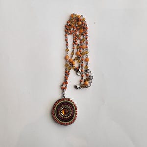 May include: A long necklace with a round pendant. The necklace is made of silver chain and orange and yellow beads. The pendant is decorated with colorful stones and beads.
