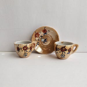 May include: Two small ceramic cups and a matching saucer with a glossy beige finish. Each piece features hand-painted red and white flowers with dark blue stems and leaves. The cups have a single handle and gold-toned rims, while the saucer has a central indentation for a cup.