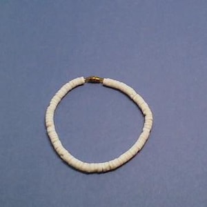 May include: White beaded bracelet with a gold-colored clasp. The bracelet is made of small, flat, round beads. The bracelet is shown on a solid blue background. The beads appear to be made of a natural material.