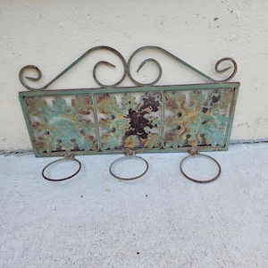 May include: A distressed metal wall planter with three circular pot holders. The rectangular planter has a turquoise and brown finish with decorative leaf cutouts. The top features scrolled metalwork.