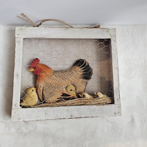 May include: A rustic, framed wall art piece featuring a hen and her chicks. The hen is brown, tan, and red, with a black tail. The chicks are yellow. The artwork is set behind wire mesh within a white-painted wooden frame.