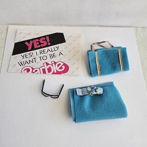 May include: A set of doll accessories including a teal felt handbag with gold trim, a matching teal felt skirt with a small blue floral embellishment, and black-framed sunglasses, alongside a card with the text YES! YES! I REALLY WANT TO BE A Barbie.
