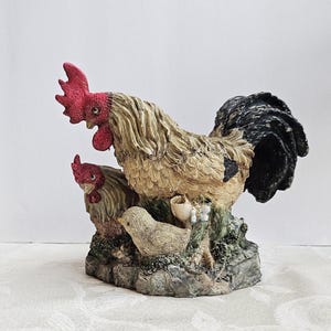 May include: A detailed figurine of a rooster, hen, and chick. The rooster has a red comb and wattle, with tan and black feathers. The hen and chick are tan. The base is a stone-like texture with green and white accents.