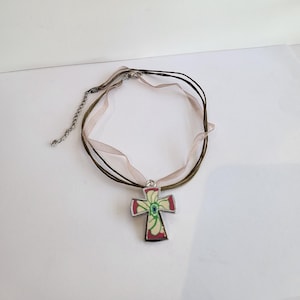 May include: A silver cross pendant with a floral design in shades of red, green, and white hangs from a brown and beige ribbon necklace.