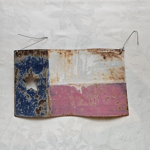 May include: A rustic metal Texas flag with a star cutout. The flag features blue, white, and red sections with a distressed, weathered appearance. Includes a wire hanger for display.