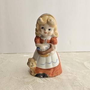 May include: A ceramic figurine of a girl in a red and white dress, holding a basket. A small cat sits at her feet. The figurine has blonde hair and is painted with delicate details. The overall theme is a vintage, collectible item.