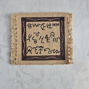 May include: Square beige coaster with a brown border and fringe. The center features black, stylized text within a brown square. The coaster is made of a woven material, likely burlap or a similar fabric.