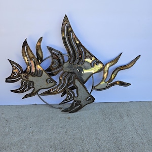 May include: Metal wall art featuring three stylized fish in shades of silver, gold, and bronze. The fish are arranged in a dynamic composition, with flowing lines and intricate details. The artwork is mounted on a circular metal frame.