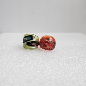 May include: Two glass rings. One ring is a light green color with dark blue, brown, and black swirls. The other ring is a reddish-orange color with black speckles. Both rings are on a white surface.