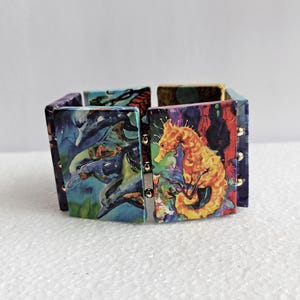 May include: A stretch bracelet composed of rectangular panels featuring colorful artistic prints. One panel displays a yellow seahorse against a dark background, while another shows blue dolphins. The panels are connected by small gold-toned metal beads.