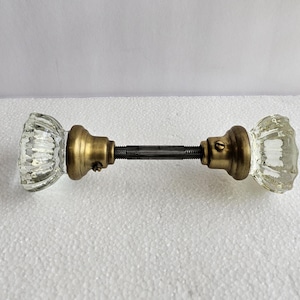 May include: A pair of clear, fluted glass door knobs connected by a central metal spindle. Each knob features a circular brass-toned base with a small set screw visible on the side, securing the glass to the threaded metal rod assembly.