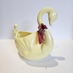May include: A pale yellow ceramic swan planter with a glossy finish. The swan features a long, curved neck adorned with a dark burgundy ribbon tied in a bow, accented by two small, dusty rose-colored fabric flowers attached to the neck.