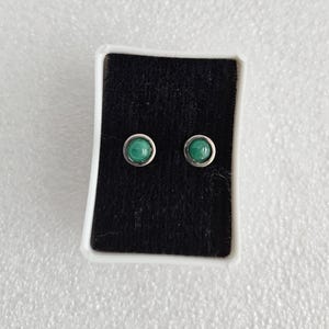 May include: A pair of stud earrings featuring round, vibrant green stones set in silver-colored metal. The earrings are displayed on a black velvet surface inside a white box, suitable for jewelry storage or presentation.