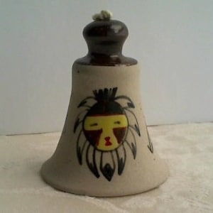 May include: A ceramic bell with a brown top and a white base. The bell is decorated with a Native American-inspired design featuring a stylized face with black hair, yellow skin, and red and black details.