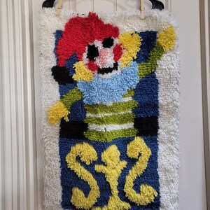 May include: A handmade, textured wall hanging featuring a colorful clown design. The clown has a red, fluffy hair, a blue ruffled collar, and a smiling face. The background is blue with yellow accents and a white, shaggy border. The artwork is hung on a black hanger.