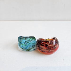 May include: Two glass rings with rounded, thick bands. One ring features a swirling pattern of light blue, dark blue, and green tones. The second ring displays a deep red and orange base with embedded gold-colored flecks throughout the glass.