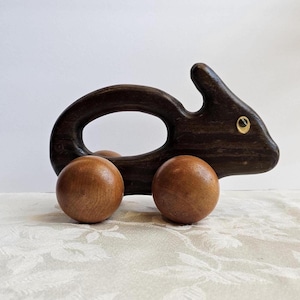 May include: A wooden toy shaped like a rabbit, with a dark brown body and two wooden wheels. The rabbit has a large oval opening in the center and a small gold-colored eye. The toy is sitting on a white floral patterned surface.