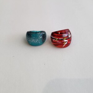 May include: Two chunky glass rings. One ring is a translucent teal blue with a speckled, glittery interior texture. The second ring is a glossy red with horizontal black stripes and small, scattered yellow dots throughout the glass surface.