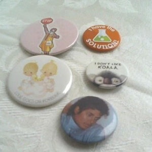 May include: Five round button pins with various designs. One features a cartoon person holding a stop sign, another has a cartoon koala with the text "I don't like koala", another has a cartoon image of two children with the text "Love Kisses Forever", another has a cartoon beaker with the text "I have the solution", and the last one has a photo of Michael Jackson.