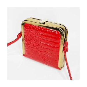 May include: A bright red crossbody bag featuring a glossy crocodile-embossed texture. The bag has a structured rectangular shape with a polished gold-tone metal frame and a matching gold-tone clasp closure at the top. It includes a thin red strap.