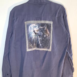 May include: A long-sleeved, button-down shirt in a muted blue hue. The back features a rectangular graphic of a wolf with a full moon in the background. The wolf is depicted in shades of gray and brown, with a detailed fur texture.