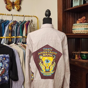 May include: A white button-down shirt with a geometric pattern. The shirt has a brown patch on the back with a graphic of an eye, hands, and the text "Trey Anastasio Band Paper Wheels Tour 2017."