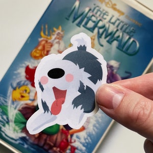 Pu&ograve; includere: Un adesivo di un cane di cartone animato bianco e nero con la lingua rossa che sporge. L'adesivo &egrave; posizionato su una custodia VHS per il film Disney "La Sirenetta".