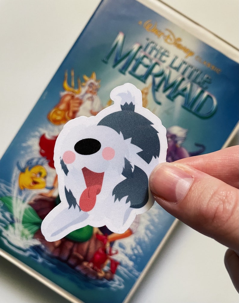 Pu&ograve; includere: Un adesivo di un cane di cartone animato bianco e nero con la lingua rossa che sporge. L'adesivo &egrave; posizionato su una custodia VHS per il film Disney "La Sirenetta".