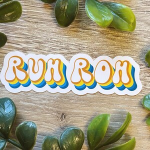 May include: A white sticker with the phrase "RUH ROH" in a retro, colorful font. The letters are outlined in blue and yellow, with a white background.