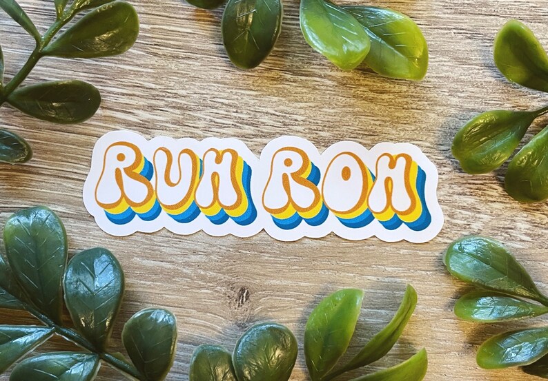 May include: A white sticker with the phrase "RUH ROH" in a retro, colorful font. The letters are outlined in blue and yellow, with a white background.