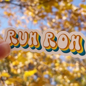 May include: A white sticker with the phrase "RUH ROH" in a retro, colorful font. The letters are outlined in yellow, blue, and orange.