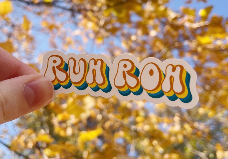 May include: A white sticker with the phrase "RUH ROH" in a retro, colorful font. The letters are outlined in yellow, blue, and orange.