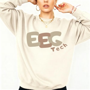 May include: Cream-colored sweatshirt with the text "EEG Tech" in a brown and tan color scheme. The sweatshirt has a crew neck and long sleeves. The text is in a bold, rounded font.
