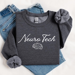 Puede incluir: Una sudadera de cuello redondo en color gris carbón oscuro con el texto blanco Neuro Tech en fuente cursiva sobre un dibujo lineal blanco de un cerebro humano. La sudadera tiene mangas largas con puños y cuello de canalé.