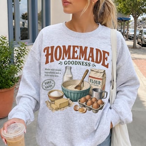 May include: A light gray crewneck sweatshirt featuring a graphic print with the text "HOMEMADE GOODNESS" and "BAKE HAPPY". The design includes illustrations of a flour bag, a mixing bowl with a wooden spoon, a milk bottle, butter, and eggs.