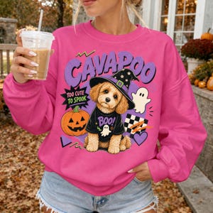 Puede incluir: Una sudadera rosa brillante con un gráfico de un perro Cavapoo que lleva un sombrero de bruja y una camiseta negra con el texto 'BOO'. El diseño incluye el texto 'CAVAPOO' y 'TOO CUTE TO SPOOK', una calabaza, un fantasma y un corazón.