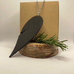 May include: A black leather pendant shaped like a wing, hanging from a silver chain. The pendant is displayed on a wooden slice with a sprig of rosemary.