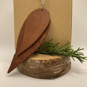 May include: A brown leather necklace with two layered feather pendants hanging from a silver chain.