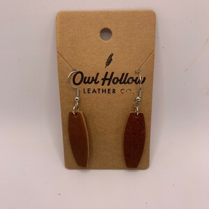 May include: A pair of brown leather earrings with silver hooks. The earrings are oval shaped and have a suede-like texture. The earrings are packaged on a brown card with the text "Owl Hollow Leather Co."
