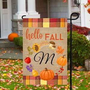 May include: A burlap garden flag with a plaid border. The flag features a wreath of fall-themed elements, including pumpkins, sunflowers, and leaves. The text "hello FALL" is printed in orange at the top of the flag, and the letter "M" is printed in black in the center of the wreath.