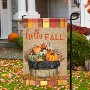 May include: A burlap garden flag with a plaid border and a basket of pumpkins, gourds, and wheat. The text "hello FALL" is written in orange on the flag.