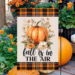 Könnte beinhalten: Eine Herbst-Gartenflagge mit einem braunen, orangen und weißen karierten Rand. Die Flagge zeigt einen großen orangefarbenen Kürbis mit einem Blumendesign und dem Text "fall is in the air".