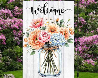 May include: A garden flag featuring the word "Welcome" in cursive script. Below is a watercolour illustration of a floral bouquet in a mason jar. The flowers are in shades of pink, orange, and blue, set against a white wooden background.