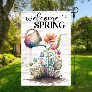 Puede incluir: Bandera de jardín con la frase "welcome SPRING". Ilustración acuarela de una regadera vertiendo agua sobre flores coloridas. El fondo es un diseño de vetas de madera blanca.