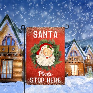 Op de afbeelding: Een rode kersttuin vlag met een wit gezicht van de kerstman in een groene krans. De tekst op de vlag luidt "SANTA Please STOP HERE".