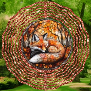 May include: A decorative wind spinner featuring a sleeping fox in a forest scene. The fox has orange and white fur, surrounded by autumn leaves. The outer ring has a red glitter design, with a green and brown background.